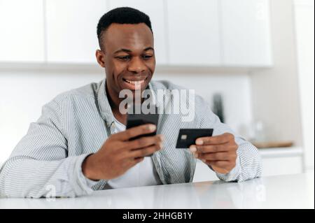 Online-Shopping. Zufriedener Schwarzer kauft von zu Hause aus in Online-Shops und Anwendungen, verwendet Smartphone und Bankkarte, um Einkäufe und Lieferungen zu bezahlen, gibt Daten ein, schaut auf die Karte, lächelt Stockfoto