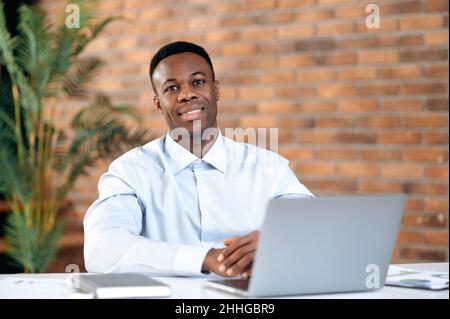 Porträt eines selbstbewussten, erfolgreichen, smarten, gutaussehenden afroamerikanischen Geschäftsmanns, ceo oder Top-Managers, sitzend im modernen Büro, mit einem Laptop zur Arbeit, in formeller Kleidung, mit Blick auf die Kamera, lächelnd Stockfoto
