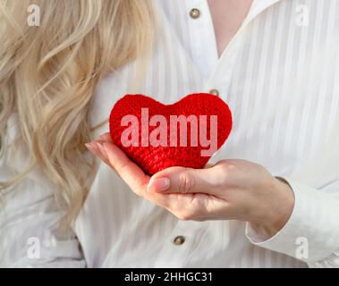 Eine blonde Frau in einer weißen Bluse hält ein rot gestricktes Herz in der Hand. Das Konzept von Pflege, Liebe und Valentinstag. Herz-Kreislauf-Erkrankungen. Wählen Sie Stockfoto