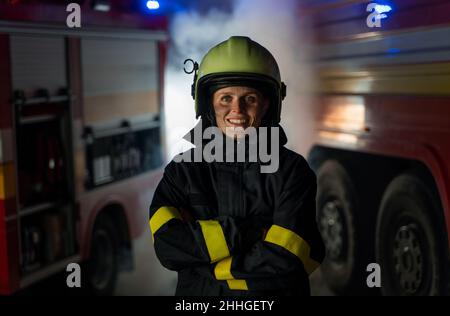 Glückliche schmutzige weibliche Feuerwehrmann Blick auf Kamera mit Feuerwehrauto im Hintergrund in der Nacht. Stockfoto