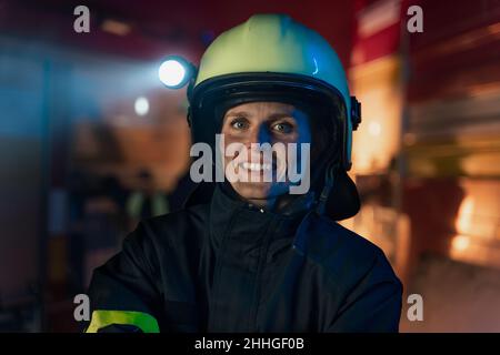 Glückliche schmutzige weibliche Feuerwehrmann Blick auf Kamera mit Feuerwehrauto im Hintergrund in der Nacht. Stockfoto