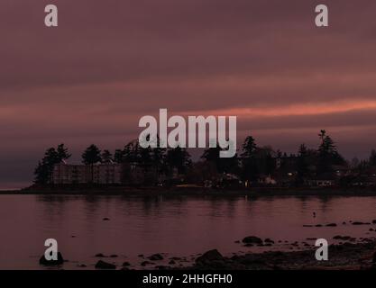 Rosa Abendlicht mit dunklen Silhouetten von Bäumen, Gebäuden mit Lichtern auf einem Landstrich am Strand, Stockfoto