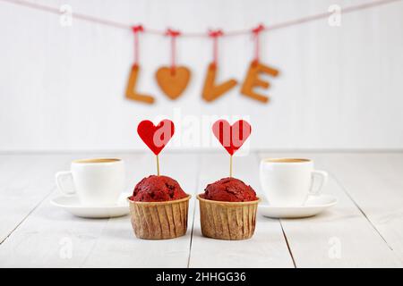 Zwei hausgemachte rote Samtcupcakes mit roten Filzherzen und zwei Tassen Kaffee auf einem weißen Holztisch. Stockfoto