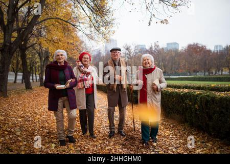 Eine Gruppe von glücklichen älteren Freunden mit Büchern, die im Herbst im Park spazieren gehen, reden und lachen Stockfoto