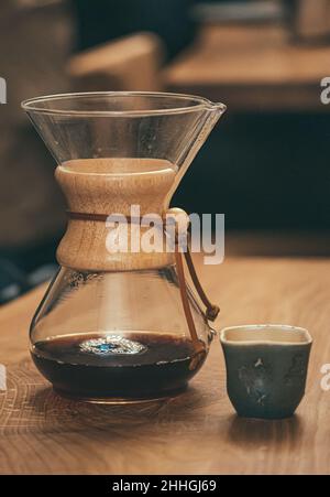 Amerikanischer Kaffee, der mit einem Papierfilter und einer chemex-Kaffeemaschine mit kleiner Keramikschale auf einem vertikalen Holztisch serviert wird Stockfoto