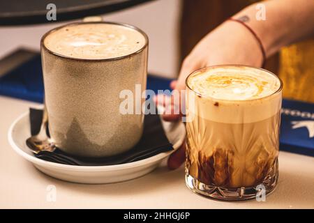 Ein paar Cappuccino mit Schaum, serviert von der Hand eines Mädchens an einer Theke, einer in einem Keramikbecher, einer in einem Trinkglas Stockfoto