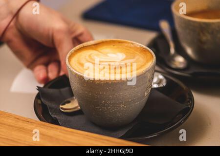 Keramikbecher oder Tasse Cappuccino mit Schaum, serviert von der Hand eines Mädchens an einer Theke Stockfoto