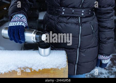 Eine Frau gießt im Winter Tee aus einer Thermoskanne auf der Straße in einen Tsakan. Hochwertige Fotos Stockfoto