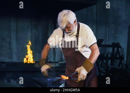 Starker reifer Mann in Schutzhandschuhen und Schürze, die geschmolzenem Stahl mit einer Zepel biegt. Kaukasischer grauhaariger Schmied, der bei der Schmiede Metall auf Amboss formt. Stockfoto