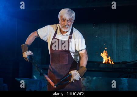 Kaukasischer Schmied, der in der Schmiede geschmolzenes Stahl mit einer industriellen Zepsie formte. Starker grauhaariger Mann mit Sicherheitsschürze und Handschuhen. Manuelles Arbeitskonzept. Stockfoto