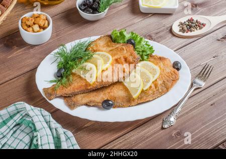 Gebratener Flunder-Fisch mit Zitrone und Kräutern auf einem weißen Teller auf dunklem Holzgrund. Stockfoto