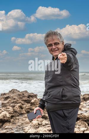 Porträt eines grauhaarigen erwachsenen Mannes mit einer Brille, die auf die Kamera zeigt, während er mit dem Meer hinter sich lächelt. Stockfoto