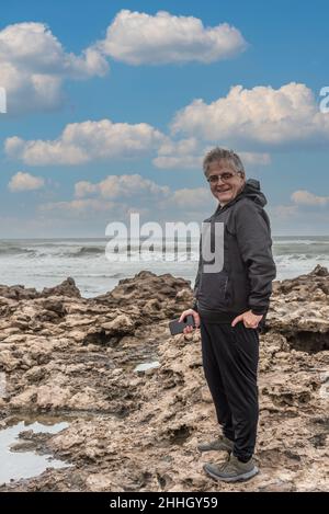Porträt eines grauhaarigen erwachsenen Mannes mit Brille, der auf die Kamera blickt, während er mit dem Meer und den Wolken hinter ihm lächelt. Stockfoto