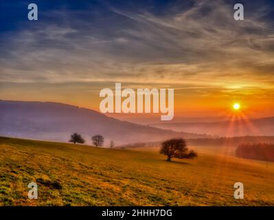 Malerische hügelige Landschaft mit einsamen Bäumen bei Sonnenuntergang, Blick auf das Tal, blauer Himmel mit hohen Wolken, Sonne. Herbstabend. Weiße Karpaten mountai Stockfoto