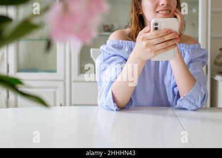 Fröhliche stilvolle Frau in blauer Bluse überprüft soziale Netzwerke im Smartphone sitzen am weißen Tisch mit Blumen auf den Vordergrund zu Hause Nahaufnahme Stockfoto