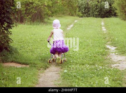 Entzückende kleine Mädchen in einem lila Pettirock gekleidet zu Fuß mit ihrer Kutsche im Sommerpark Stockfoto