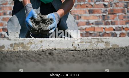 Nahaufnahme von Menschenhänden in Arbeitshandschuhen mit Sand-Zement-Mischung für Estrich. Männlicher Arbeiter, der auf der Baustelle Estrich-Material vorbereitete, während er in der Nähe der Estrich-Schiene kniete. Stockfoto