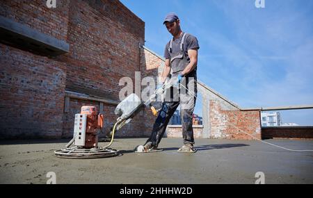 Seitenansicht des jungen Arbeiters in Schutzhandschuhen, die Betonplatten mit einer Spezialmaschine ausgleichen. Konzept der Prozesskonstruktion neues modernes Haus mit speziellen Ausrüstungen im Freien. Stockfoto