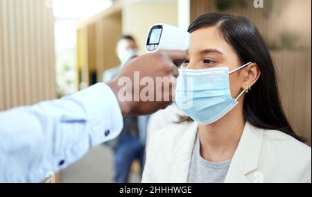 Lass uns deine Temperatur messen. Ausgeschnittene Aufnahme einer attraktiven jungen Geschäftsfrau, die eine Maske trägt und ihre Temperatur während des Stehens an der abnimmt Stockfoto