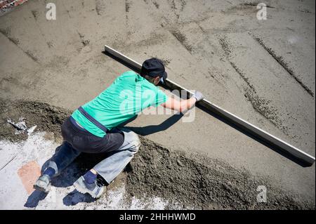 Draufsicht auf einen männlichen Arbeiter, der auf der Baustelle Estrichschienen auf den mit Sand-Zement-Gemisch überzogenen Boden legte. Man nivellieren Oberfläche mit gerader Kante, während Estrichboden im Freien im neuen Gebäude. Stockfoto