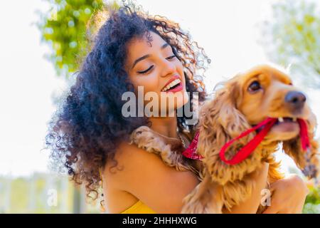 Schöne Mixrd-Rennfrau mit Make-up und lockiger Frisur, die an sonnigen Tagen auf der Straße mit ihrem amerikanischen Cockerhund Cite Gold glückliche Zeiten verbringt Stockfoto