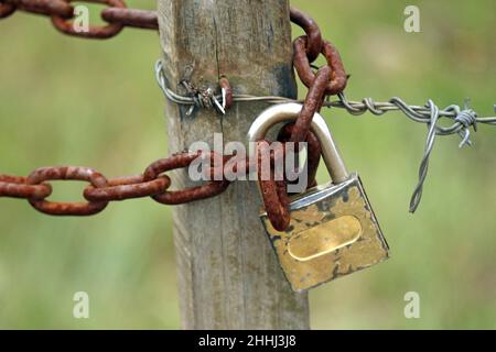 Ein kleiner Holzmast, der durch Stacheldraht aufgehängt ist, eine rostige Kette und ein kleines Vorhängeschloss. Im einfachen Leben auf dem Land gibt es keine Mauern. Stockfoto