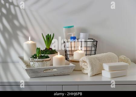 Badezimmereinrichtung. Ungeblasene Hyazinthen mit brennenden Kerzen auf einem hölzernen Vintage-Tablett. Palmenbaum-Schatten an der Wand. Shampoos und Creme in einem Metallkorb. R Stockfoto