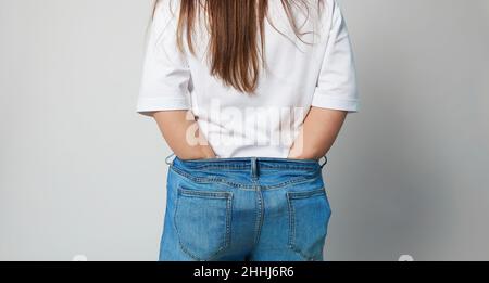 Ein Mädchen in blauen Jeans und einem weißen T-Shirt von hinten, das ihre Hände in Jeans legt, die für sie groß geworden sind, zeigt, wie sie an Gewicht verloren hat. Fragment Stockfoto