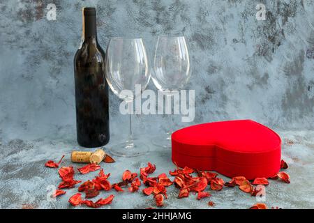 Zutaten zum Valentinstag auf blauem Hintergrund: Rotwein-Flasche, zwei Weingläser, rotes herzförmiges Geschenkkarton und trockene Rosenblätter. Seiten- oder Profilansicht. Stockfoto