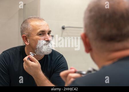 Gut aussehender reifer Mann mit grauen Haaren versucht, seinen Bart selbst in einem Badezimmer abzuschneiden Stockfoto