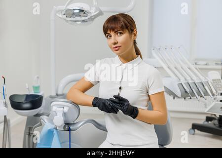 Frau Zahnärztin in weißen Uniform und Handschuhe sitzen in Stomatologie Schrank unter modernen zahnmedizinischen Geräten. Porträt einer Ärztin - professioneller Stomatologe, Kieferorthopäde. Stockfoto