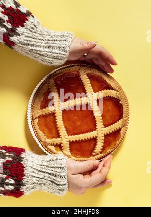 italienische Krostatkuchen mit Aprikosenmarmelade auf gelbem Hintergrund und weiblichen Händen in einem warmen Wollpullover mit Ornament Stockfoto