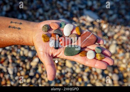 Konzeptuelles Foto einer Sommerstimmung. Männliche Hand mit kleinen bunten Kieselsteinen auf der Handfläche. Kristalle Meeresfrüchtevererze. Glücklicher Urlaub im Beach Resort. Stockfoto