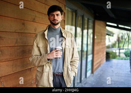 Ernsthafter Mann, der mit der Hand in der Jackentasche und einer Tasse Getränk zum Mitnehmen steht und die Kamera anschaut Stockfoto