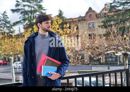 Nachdenklicher männlicher Student, der Bücher für Studien auf Gehwegen im Stadtpark trägt Stockfoto