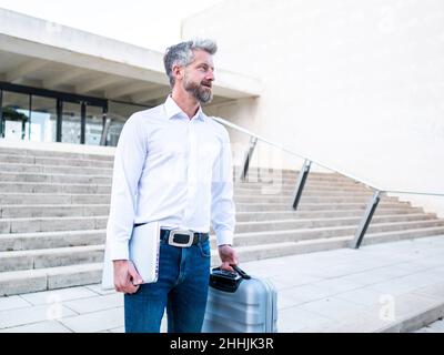Seriöse bärtige männliche Unternehmerin mit modernem Netbook und Gepäck, die während der Reise in die Stadt in der Nähe des Gebäudes nach unten läuft Stockfoto