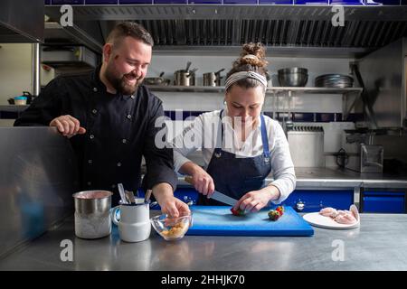 Glücklicher bärtiger Mann, der lächelte und eine Schüssel mit Krümel nahm, während eine Kollegin während der Dessertvorbereitung in der Restaurantküche Erdbeeren Schnitt Stockfoto