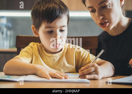 Junge macht seine Hausaufgaben mit seiner Mutter sitzt am Schreibtisch, zu Hause, er schreibt auf Copybook. Entwicklung von Kindern. Stockfoto