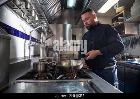 Ein bärtiger Mann in dunkler Uniform, der eine Bratpfanne auf die Kochplatte legt, während er in der modernen Restaurantküche Gericht zubereitet Stockfoto