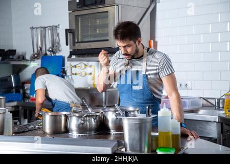 Fokussierter junger bärtiger ethnischer Koch in legerer Kleidung und Schürze, der während der Arbeit Essen mit einem Löffel am Herd mit verschiedenen Metallkochköpfen verkostet Stockfoto