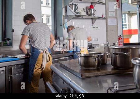 Rückansicht des jungen hispanischen männlichen Kochs in legerer Kleidung und Schürze, der Süßkartoffeln schneidet, während er in der Restaurantküche in der Nähe eines Kollegen arbeitet Stockfoto