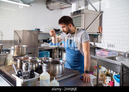 Fokussierter junger bärtiger ethnischer Koch in legerer Kleidung und Schürze, der während der Arbeit Essen mit einem Löffel am Herd mit verschiedenen Metallkochköpfen verkostet Stockfoto
