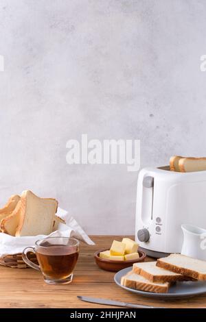 Tasse Tee mit Butterwürfeln und Scheiben frisches Brot auf Holztisch in der Nähe des modernen Toasters in der Küche in sonnigen Morgen platziert Stockfoto