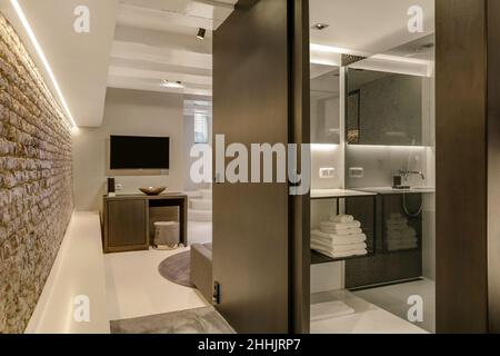 Interieur der modernen Wohnung mit TV an der weißen Wand über dem Tisch und Handtücher auf Regalen im Badezimmer mit offener Tür Stockfoto