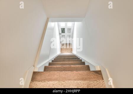 Von oben eine lange Treppe mit braunen Stufen, die zur Tür in engem Raum mit weißen Wänden führt Stockfoto