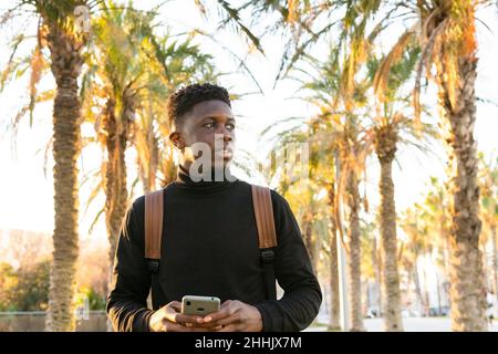 Afroamerikanischer Kerl, der auf dem Handy in der Nähe von grünen Palmen stöbert Stockfoto