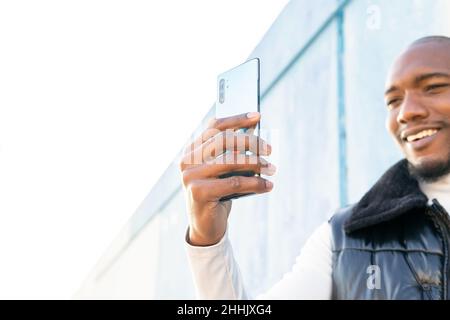 Glücklicher, kahler afroamerikanischer Mann, der mit dem Finger auf den Bildschirm des Smartphones zeigt, während er in der Nähe einer Mauer in der Stadt ein Selbstporträt aufnahm Stockfoto