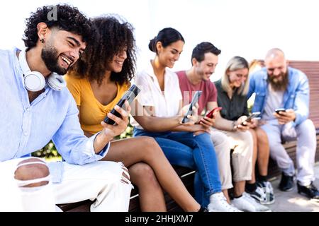 Fröhliche junge Gruppe von Menschen, die zusammen mit dem Handy im Freien sitzen Stockfoto