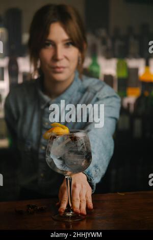 Die Barkeeperin mit weichem Fokus blickt auf die Kamera, während sie Weinglas mit Gin und Tonic-Cocktail serviert, der mit orangefarbener Schale auf der Holztheke in der Bar dekoriert ist Stockfoto