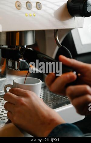 Bei der Arbeit in einem hellen Café wird ein nicht erkennbarer Barista mit einer weißen Tasse für den heißen Espresso zubereitet, während eine moderne Kaffeemaschine mit Portafilter verwendet wird Stockfoto